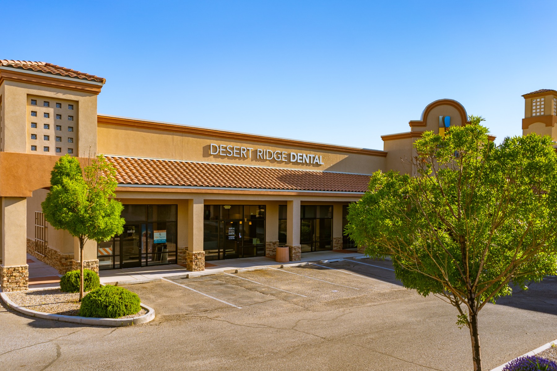 A wide-angle view of the Desert Ridge Dental office building, showcasing its exterior. The building features a tan stucco facade with stone accents and a tile roof, typical of southwestern architecture. The name "Desert Ridge Dental" is prominently displayed above the entrance. The surrounding area includes neatly landscaped greenery with small trees and shrubs.