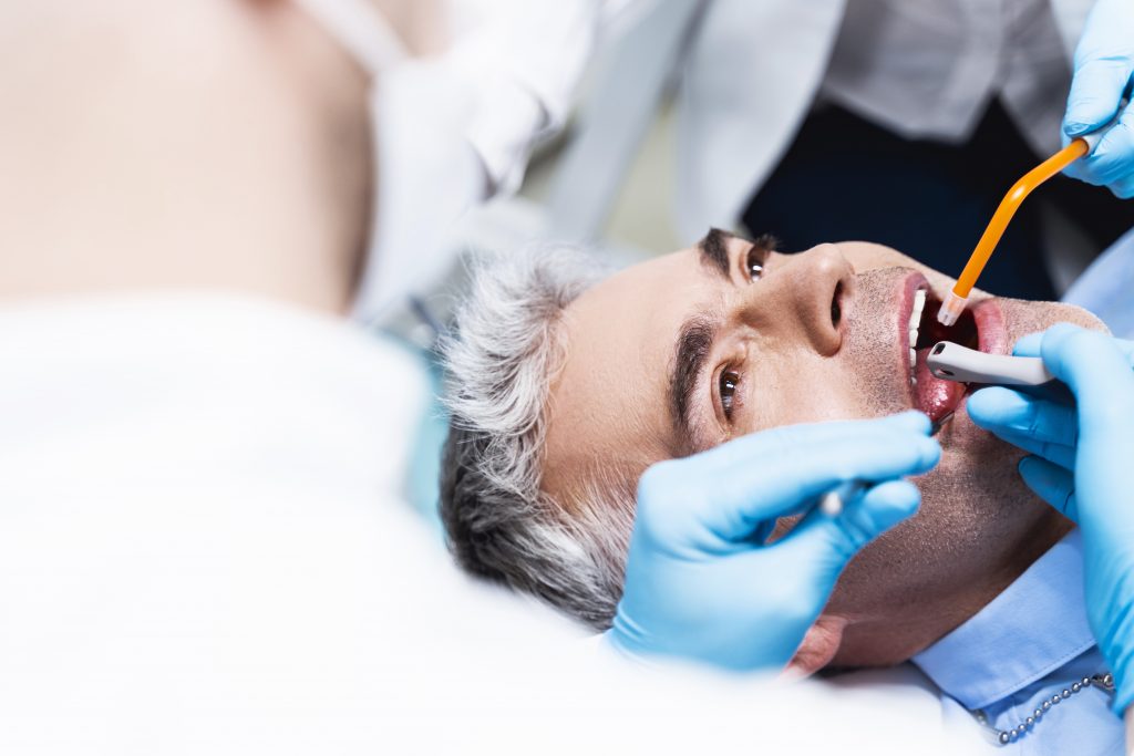 a man lying back in a dental chair during a dental procedure. The dentist and dental assistant, both wearing blue gloves, are working in the man's mouth. One is using a dental tool, while the other holds a suction device. The man appears calm and cooperative, looking upward as the dental professionals perform the procedure.