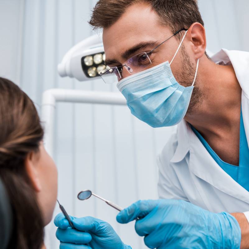 Dr. Mink, in black scrubs is pointing to an X-ray image displayed on a monitor. The X-ray shows a detailed view of a patient's teeth and jaw. The dentist is explaining something to a patient.